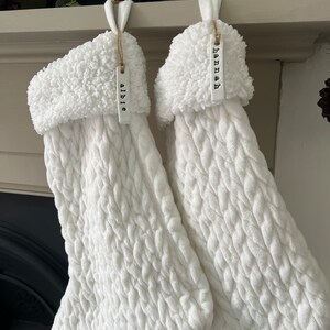 May include: Two white knitted stockings with fluffy white cuffs hanging from a fireplace mantel. The stockings have personalized tags that read "Abbie" and "Hannah".
