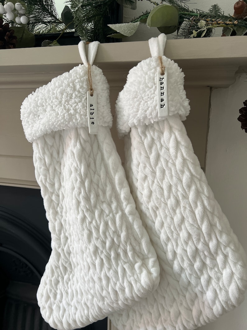 May include: Two white knitted stockings with fluffy white cuffs hanging from a fireplace mantel. The stockings have personalized tags that read "Abbie" and "Hannah".
