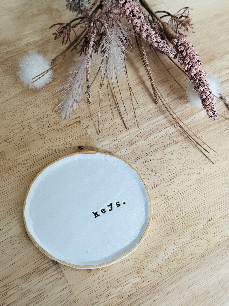 May include: White ceramic dish with a gold rim and the word "keys." written in black lettering. The dish is on a light wood surface.