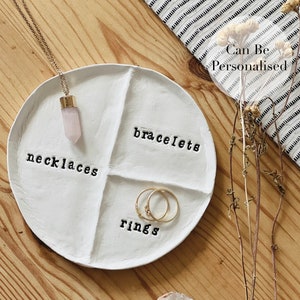 Könnte beinhalten: Ein weißes, rundes Schmucktablett mit vier Abschnitten, beschriftet mit "Halsketten", "Armbänder" und "Ringe". Eine goldene Halskette mit einem rosa Kristallanhänger und zwei goldene Ringe liegen auf dem Tablett. Der Text "Can Be Personalised" ist sichtbar.