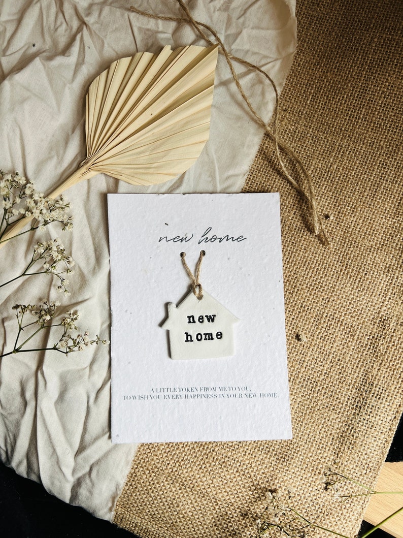 May include: A white card with the text "new home" written on it. A small white ceramic house charm with the text "new home" is attached to the card with twine.