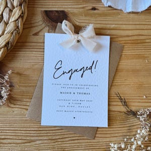 Puede incluir: Una tarjeta de anuncio de compromiso azul claro con un lazo de cinta blanco y el texto "Engaged!" en escritura negra. La tarjeta también incluye el texto "Please join us in celebrating the engagement of Maisie & Thomas" y la fecha y hora del evento.