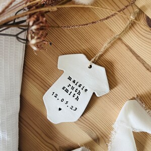 May include: White ceramic baby onesie ornament with the text "Maisie Ruth Smith 12.05.23" in black lettering.