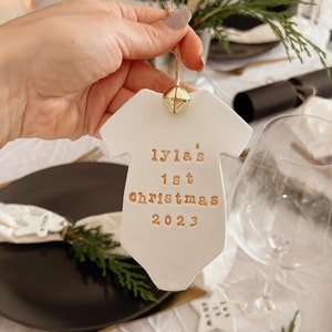 Puede incluir: Adorno de cer&aacute;mica blanco con forma de body de beb&eacute; con texto dorado "Lyla's 1st Christmas 2023". El adorno tiene una peque&ntilde;a campana dorada. Decoraci&oacute;n festiva navide&ntilde;a.