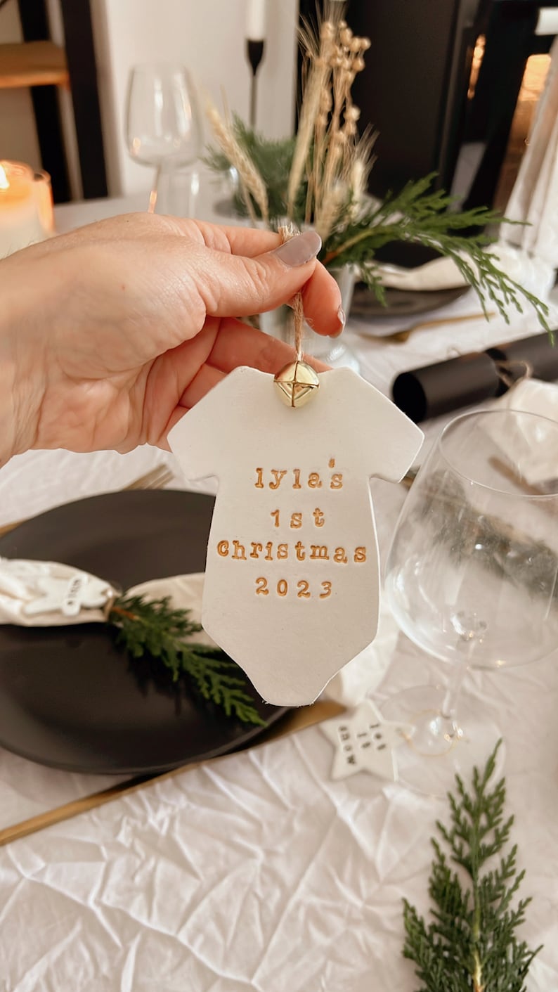 Puede incluir: Adorno de cer&aacute;mica blanco con forma de body de beb&eacute; con texto dorado "Lyla's 1st Christmas 2023". El adorno tiene una peque&ntilde;a campana dorada. Decoraci&oacute;n festiva navide&ntilde;a.
