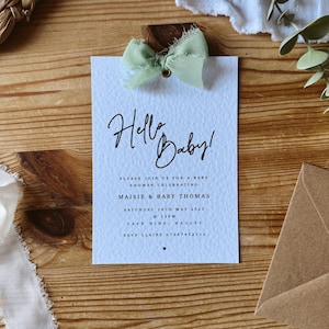 May include: A light blue baby shower invitation with a white background and a green bow. The invitation says "Hello Baby!" and includes the details for a baby shower for Maisie & Baby Thomas on Saturday, May 18th, 2025 at 12pm at Cafe Nine, Hauley. RSVP to Claire at 07987452312.