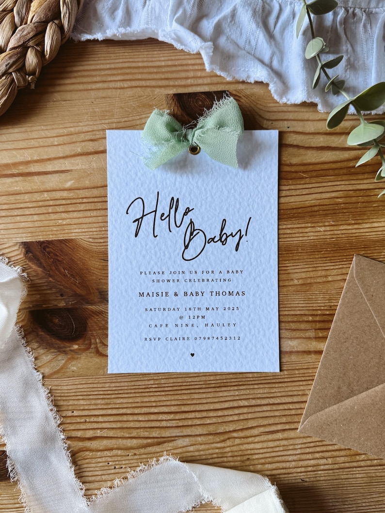May include: A light blue baby shower invitation with a white background and a green bow. The invitation says "Hello Baby!" and includes the details for a baby shower for Maisie & Baby Thomas on Saturday, May 18th, 2025 at 12pm at Cafe Nine, Hauley. RSVP to Claire at 07987452312.