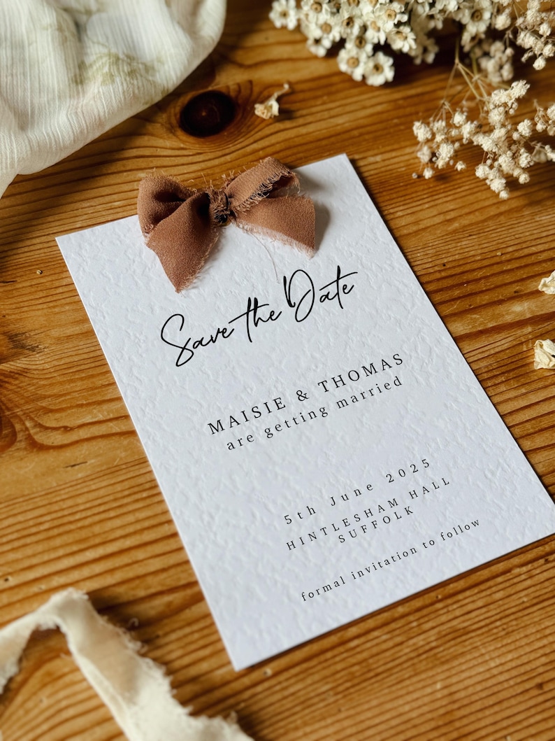 May include: Save the Date card with a brown ribbon bow. The card is white with black text that reads "Save the Date", "Maisie & Thomas", "are getting married", "5th June 2025", "Hintlesham Hall", "Suffolk", and "formal invitation to follow".