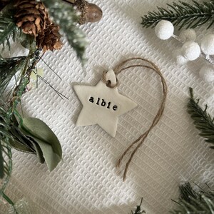 May include: A white star-shaped Christmas ornament with the name "albie" stamped in black letters. The ornament hangs from a natural twine string. The background includes pine needles, pine cones, and white berries.