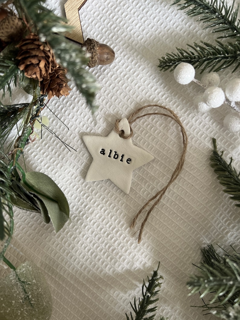 May include: A white star-shaped Christmas ornament with the name "albie" stamped in black letters. The ornament hangs from a natural twine string. The background includes pine needles, pine cones, and white berries.