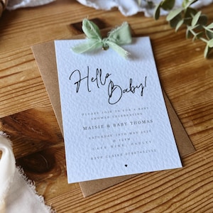 May include: A white baby shower invitation with a green ribbon. The invitation reads "Hello Baby!" and includes the details for a baby shower celebrating Maisie & Baby Thomas on Saturday, May 18th, 2025 at 12pm at Cafe Nine, Hauley. RSVP to Claire at 07987452312.