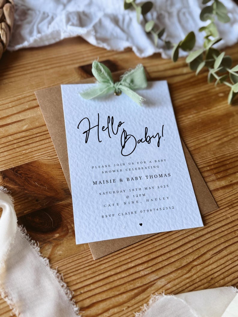 May include: A white baby shower invitation with a green ribbon. The invitation reads "Hello Baby!" and includes the details for a baby shower celebrating Maisie & Baby Thomas on Saturday, May 18th, 2025 at 12pm at Cafe Nine, Hauley. RSVP to Claire at 07987452312.