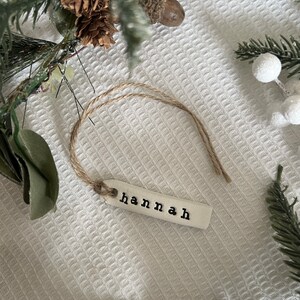 May include: A white ceramic ornament with the name "Hannah" written in black lettering. The ornament is attached to a piece of twine.
