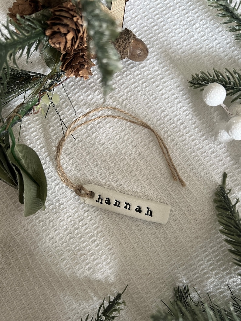 May include: A white ceramic ornament with the name "Hannah" written in black lettering. The ornament is attached to a piece of twine.