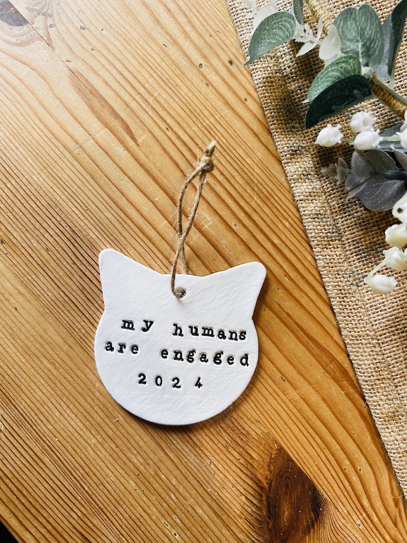May include: White ceramic cat-shaped ornament with the stamped text "my humans are engaged 2024". The ornament hangs from a natural twine string. The background is a wooden surface with a burlap cloth and greenery.