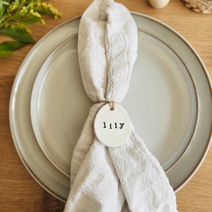 May include: A white ceramic place card with the name "Lilly" written in black lettering. The place card is tied to a beige linen napkin with twine.