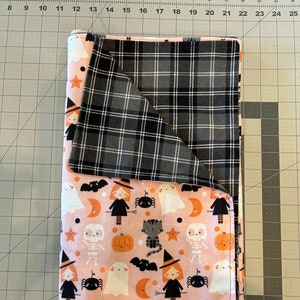 May include: Reversible Halloween-themed blanket. One side features a pink background with witch, ghost, cat, and pumpkin patterns. The other side has a black, white, and gray plaid design. The blanket is folded.