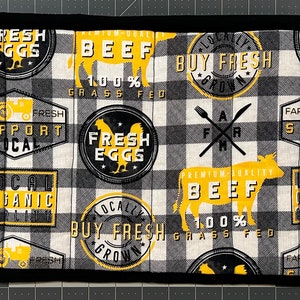 May include: A rectangular, quilted placemat with a black border. The main pattern is a black and white checkered design with yellow and black farm-themed graphics, including cows, chickens, and text that reads "Fresh Eggs" and "Beef".