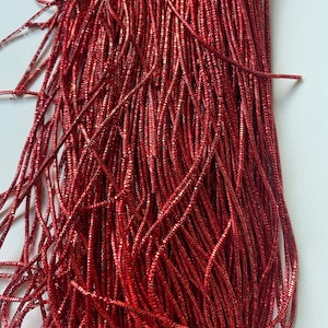 May include: Close-up of a bundle of shiny, red metallic fringe. The fringe is made of many thin, parallel strands, creating a textured and reflective surface. The strands are slightly wavy and appear to be made of a flexible material.