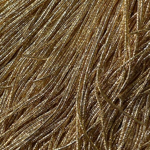 May include: A close-up of a gold metallic fringe fabric. The fringe is made of thin, shiny threads that are woven together to create a shimmering effect.
