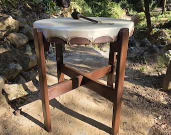 May include: A round drum with a light beige skin head stretched over a dark brown wooden frame. The drum is supported by a dark brown wooden stand featuring four legs and a cross-brace structure. A small wooden mallet with a black head rests on top.