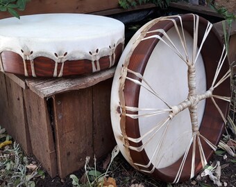 May include: Two round frame drums with dark brown wooden rims and light-colored rawhide heads. One drum sits horizontally, showing the laced edge, while the other stands vertically, displaying a central cross-lacing pattern made of thin strips.
