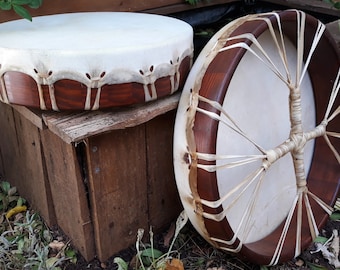 May include: Two wooden framed drums with white leather drumheads and brown leather lacing. The drums are sitting on a wooden crate.