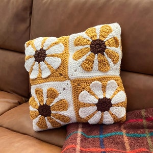 May include: A square crochet pillow with four daisy flower designs. Each flower has white petals, a brown center, and is set against a white and mustard yellow background. A brown plaid blanket is draped below the pillow on a brown leather couch.