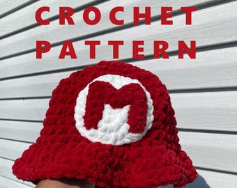 Könnte beinhalten: Ein roter Häkelhut mit einem weißen "M" Emblem. Der Hut ist aus weichem, strukturiertem Garn gefertigt und hat eine breite Krempe. Die Worte "CROCHET PATTERN" stehen in roten Blockbuchstaben über dem Hut.