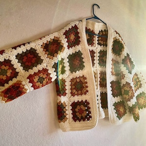 May include: A crocheted cardigan with a white background and colorful squares in shades of brown, green, and orange. The cardigan is hanging on a hanger.