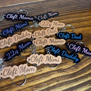 May include: A collection of keychains with various names and phrases, including "Cleft Mama," "Cleft Dad," and "Cleft Mimi." The keychains feature silver rings and chains. The text is in black, blue, and pink.
