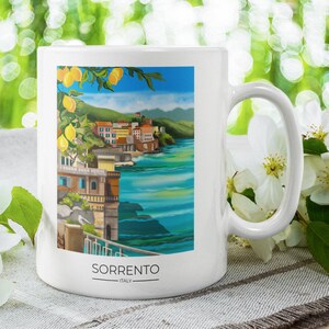 Op de afbeelding: Witte keramische mok met een kleurrijke illustratie van Sorrento, Itali&euml;, met gebouwen, de zee en citroenen. Het woord "SORRENTO" staat onderaan gedrukt. De mok heeft een gebogen oor en is omgeven door witte bloemen.