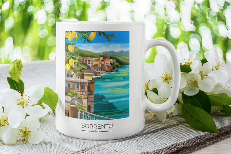Op de afbeelding: Witte keramische mok met een kleurrijke illustratie van Sorrento, Itali&euml;, met gebouwen, de zee en citroenen. Het woord "SORRENTO" staat onderaan gedrukt. De mok heeft een gebogen oor en is omgeven door witte bloemen.