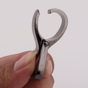 May include: A silver-toned metal clip with a rounded opening. The clip is designed to hold items together.