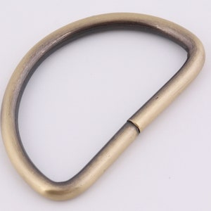 May include: A gold-toned metal D-ring with a smooth finish. The ring is open on one side and has a slightly rounded shape.