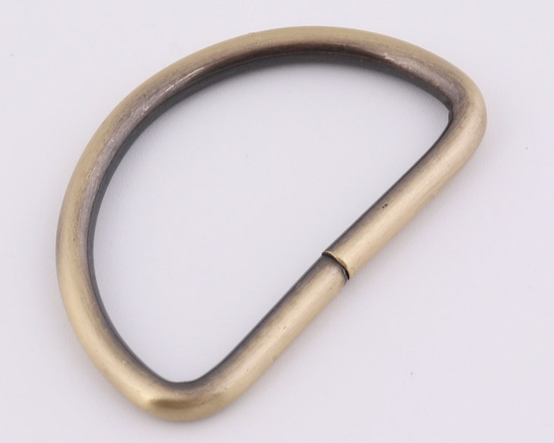 May include: A gold-toned metal D-ring with a smooth finish. The ring is open on one side and has a slightly rounded shape.