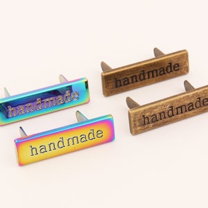 May include: Four metal tags with the word "handmade" engraved on them. Two tags are a rainbow iridescent color and two tags are a bronze color.