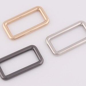 Könnte beinhalten: Drei rechteckige Metallringe in Gold, Silber und Schwarz.