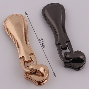 May include: Two zipper pulls, one gold and one black, with a decorative design. Each pull measures 51 mm in length.