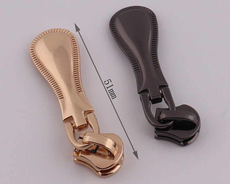 May include: Two zipper pulls, one gold and one black, with a decorative design. Each pull measures 51 mm in length.
