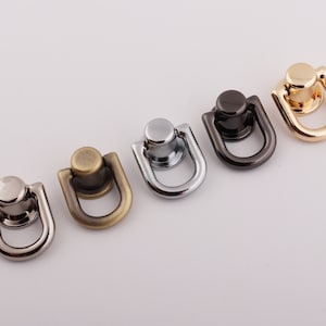 May include: An assortment of five metal D-ring hardware pieces in various finishes: silver, antique brass, chrome, black, and gold. Each D-ring has a circular top and a U-shaped base, suitable for bag making or other craft projects.