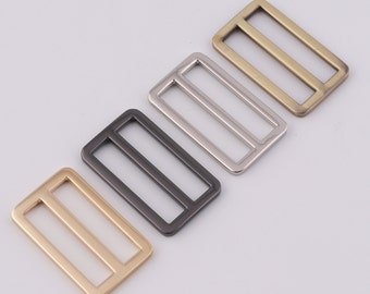 May include: Four metal slider buckles in different finishes: gold, silver, black, and antique gold. The buckles are rectangular with a single, horizontal slot in the center.