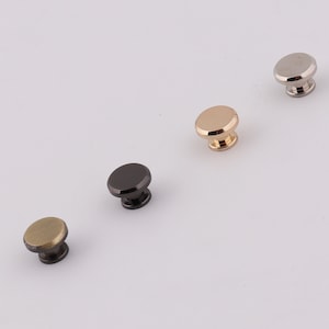 May include: Four metal knobs in different finishes: gold, silver, black, and brass. The knobs are round with a raised center and a small stem on the bottom.