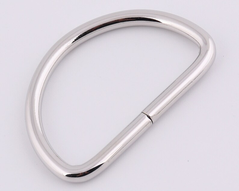 May include: A silver metal D-ring with a smooth finish. The ring is open on one side and has a closed loop on the other.