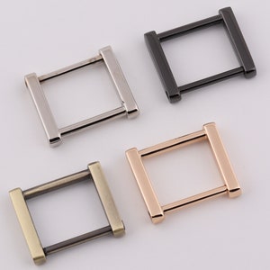 Könnte beinhalten: Vier D-Ringe aus Metall in verschiedenen Ausführungen: Silber, Schwarz, Gold und Antikmessing. Die Ringe sind quadratisch mit abgerundeten Ecken.