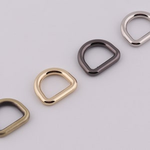 Op de afbeelding: Vier metalen D-ringen in verschillende afwerkingen: goud, zilver, zwart en brons. Deze ringen worden vaak gebruikt om banden, kettingen of andere accessoires aan te bevestigen.