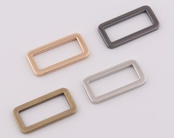 Puede incluir: Cuatro anillos rectangulares de metal en diferentes acabados: dorado, plomo, plateado y latón antiguo. Estas piezas de ferretería son adecuadas para la fabricación de bolsos, cinturones u otros proyectos de artesanía. Los anillos se muestran sobre un fondo blanco.
