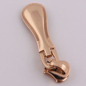 May include: A gold-colored zipper pull with a decorative top and a functional slider.