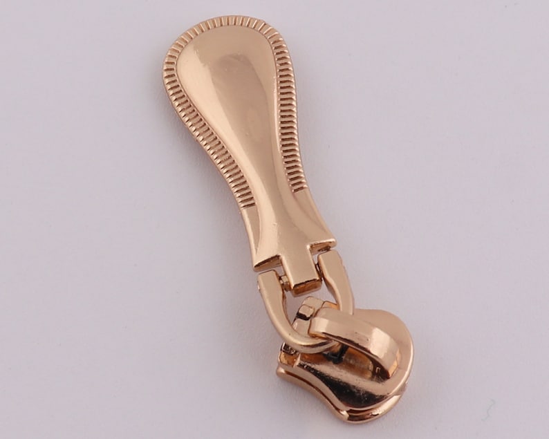May include: A gold-colored zipper pull with a decorative top and a functional slider.