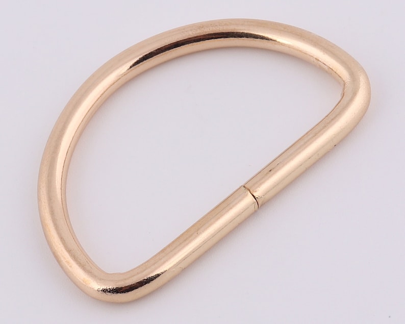 May include: A gold-colored metal D-ring, a type of hardware used for attaching straps, chains, or other accessories.